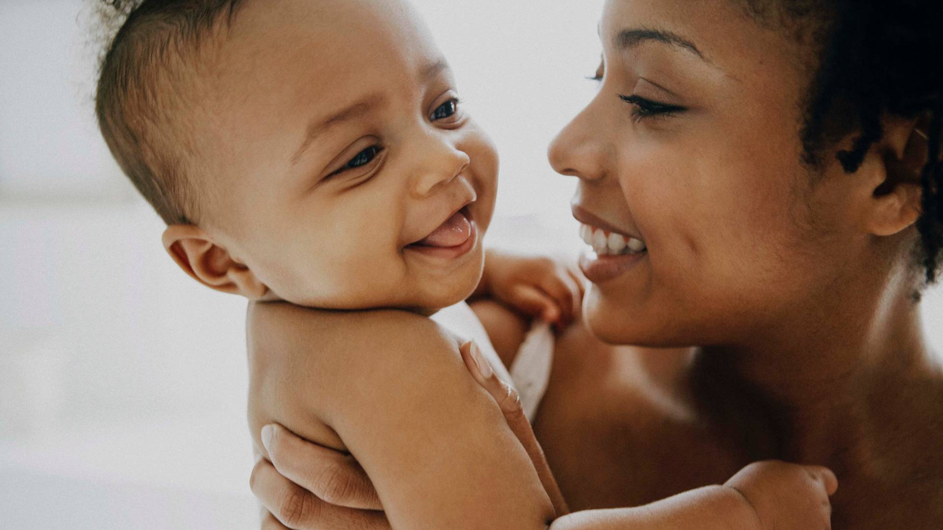 A smiling woman holds a happy baby who is looking at her and sticking out their tongue. Both appear joyful and are facing each other closely in a tender, bright setting.