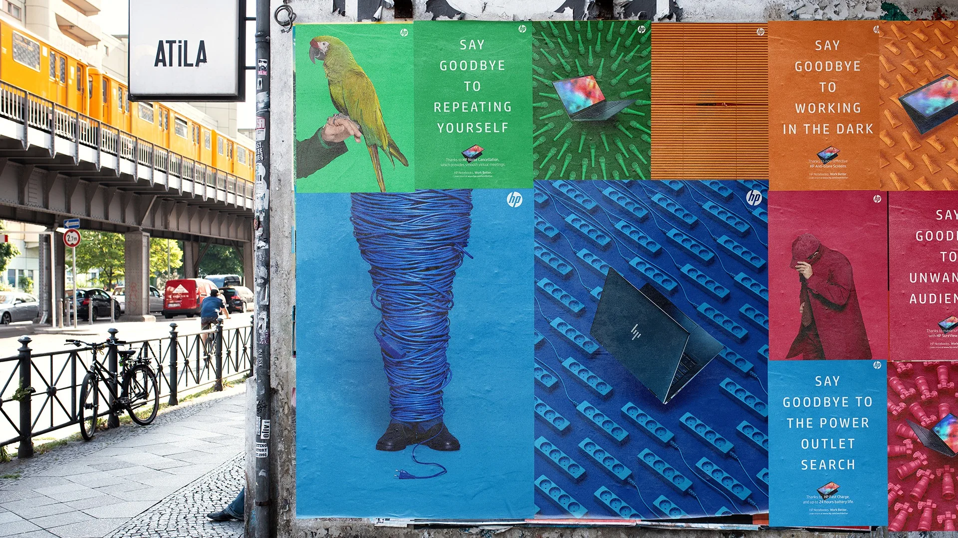 A city street scene with a wall covered in colorful HP laptop ads, each with quirky images and phrases like Say goodbye to repeating yourself and Say goodbye to working in the dark. A train passes on the left.