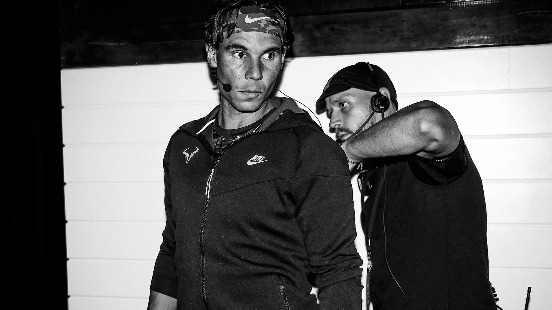 A man in sportswear with a headset stands in the foreground, while another man in dark clothing adjusts or checks his audio equipment. The image is in black and white.