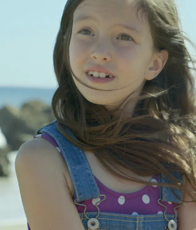 A young girl with long brown hair, wearing a purple polka dot shirt and denim overalls, looks up with a thoughtful expression. She is outdoors near a beach, with rocks and the ocean in the background.