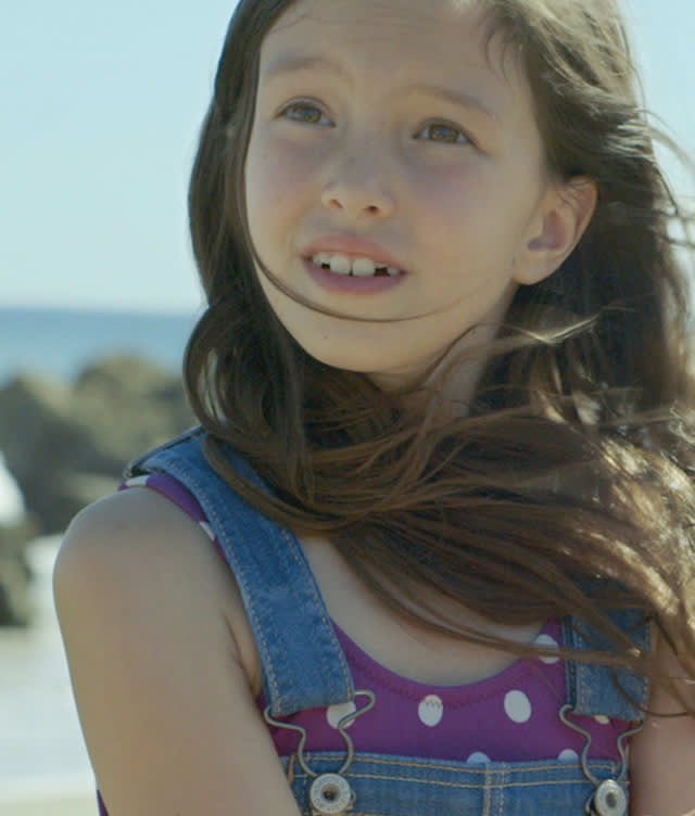 A young girl with long brown hair, wearing a purple polka dot shirt and denim overalls, looks up with a thoughtful expression. She is outdoors near a beach, with rocks and the ocean in the background.