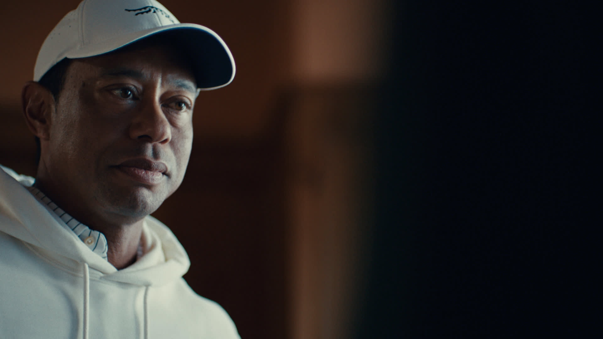 Tiger Woods wearing a white cap and white hoodie looks thoughtfully into the distance, with soft indoor lighting illuminating his face.
