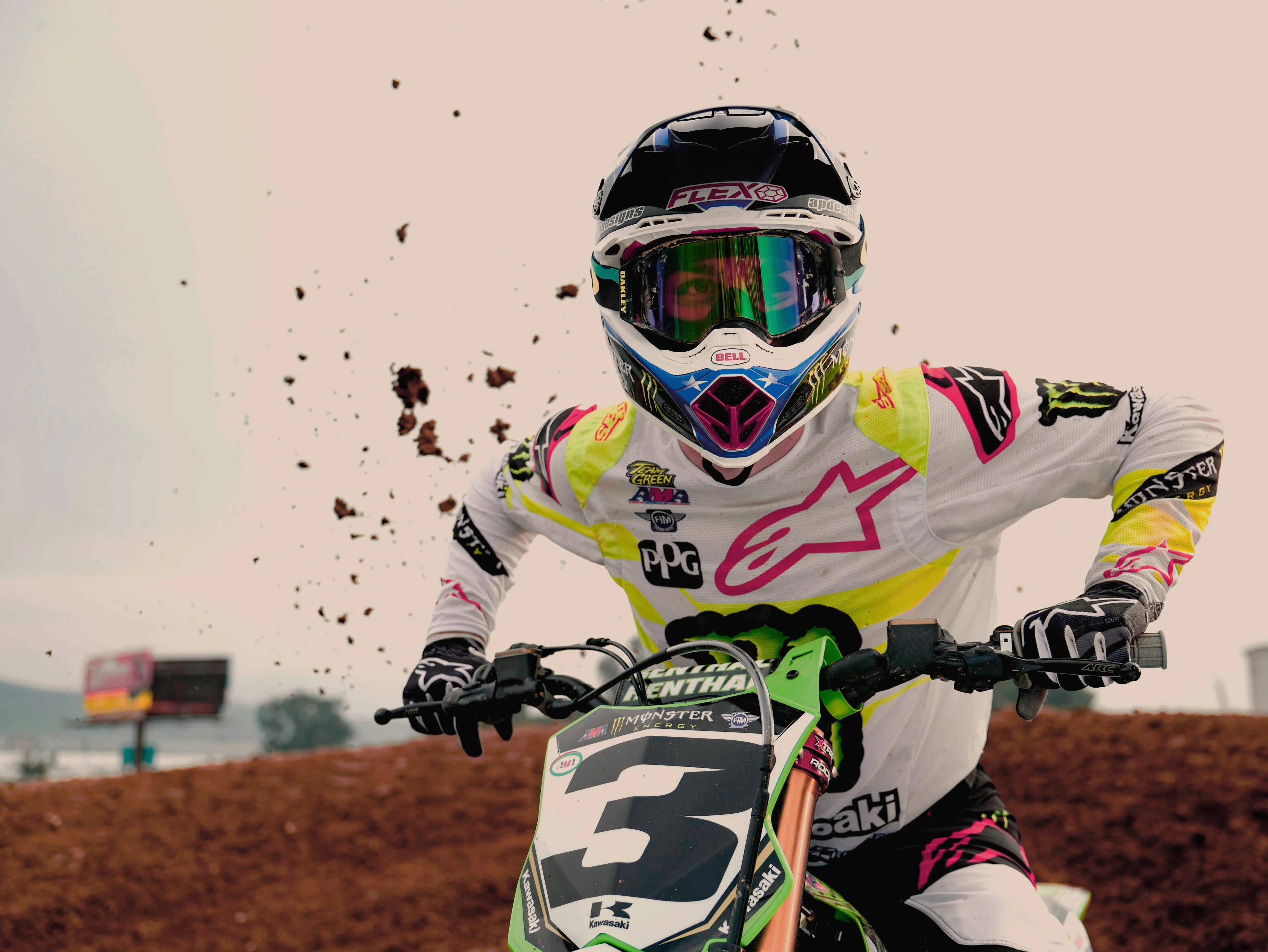 A motocross rider wearing a white and neon yellow suit and helmet rides a dirt bike with the number 3, kicking up dirt in dramatic action on a racing track.