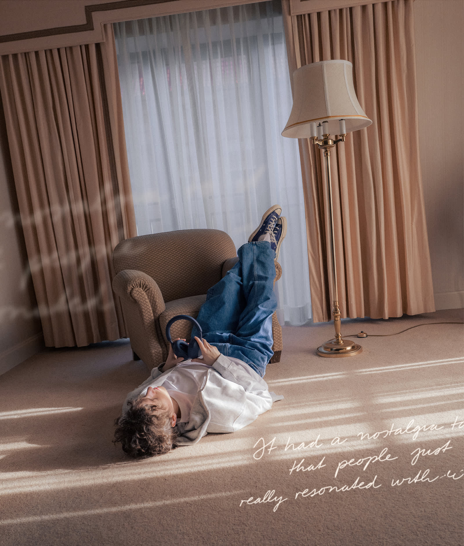 A person in jeans and a white shirt lies on the floor with legs resting on an armchair, holding headphones. The room has beige curtains, a floor lamp, and carpet. Handwritten text appears in the bottom right corner.