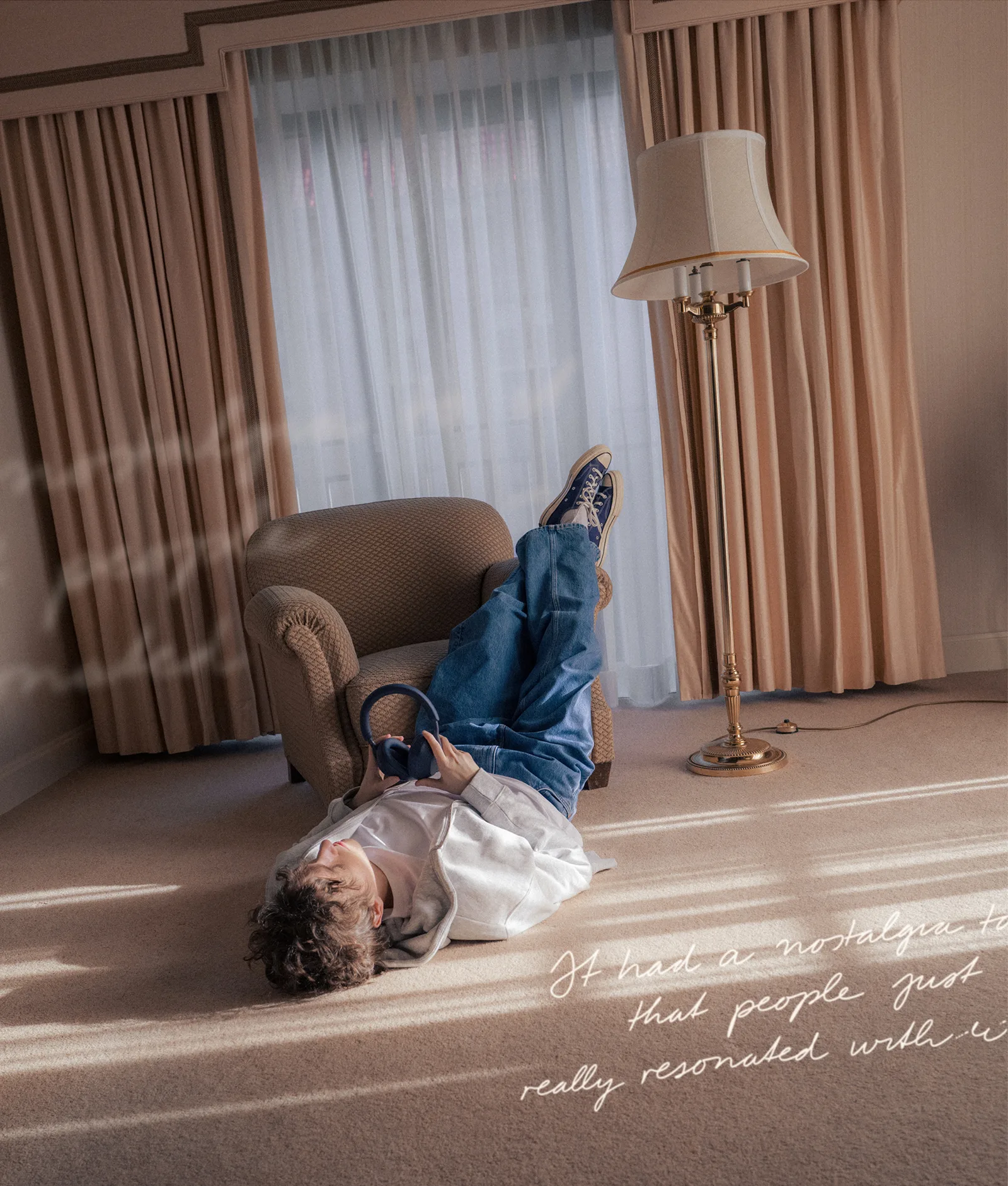 A person in jeans and a white shirt lies on the floor with legs resting on an armchair, holding headphones. The room has beige curtains, a floor lamp, and carpet. Handwritten text appears in the bottom right corner.
