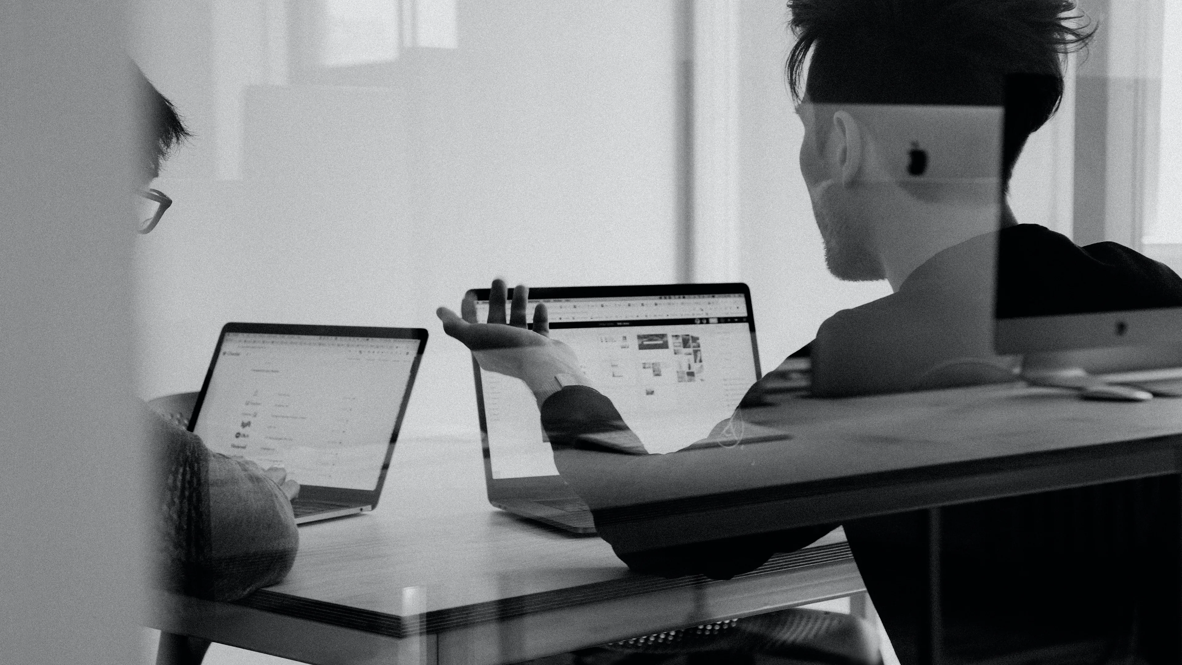 Two people sit at a desk working on laptops, discussing something on their screens. The scene is shot through glass, creating faint reflections and a collaborative office atmosphere.