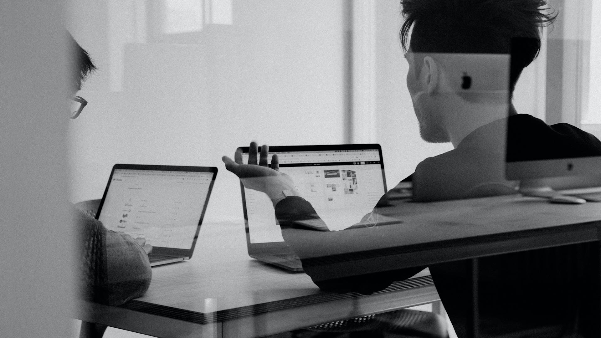 Two people sit at a desk working on laptops, discussing something on their screens. The scene is shot through glass, creating faint reflections and a collaborative office atmosphere.