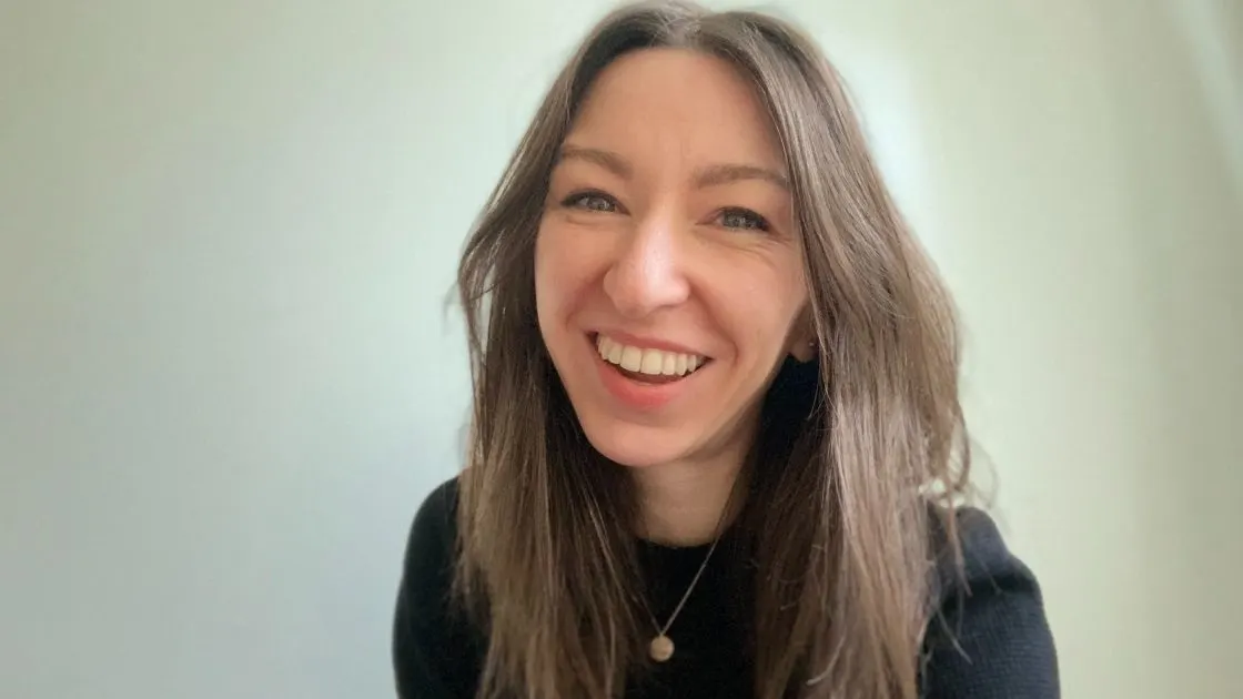 A woman with long brown hair, wearing a black top and a necklace, smiles brightly at the camera against a plain light background.
