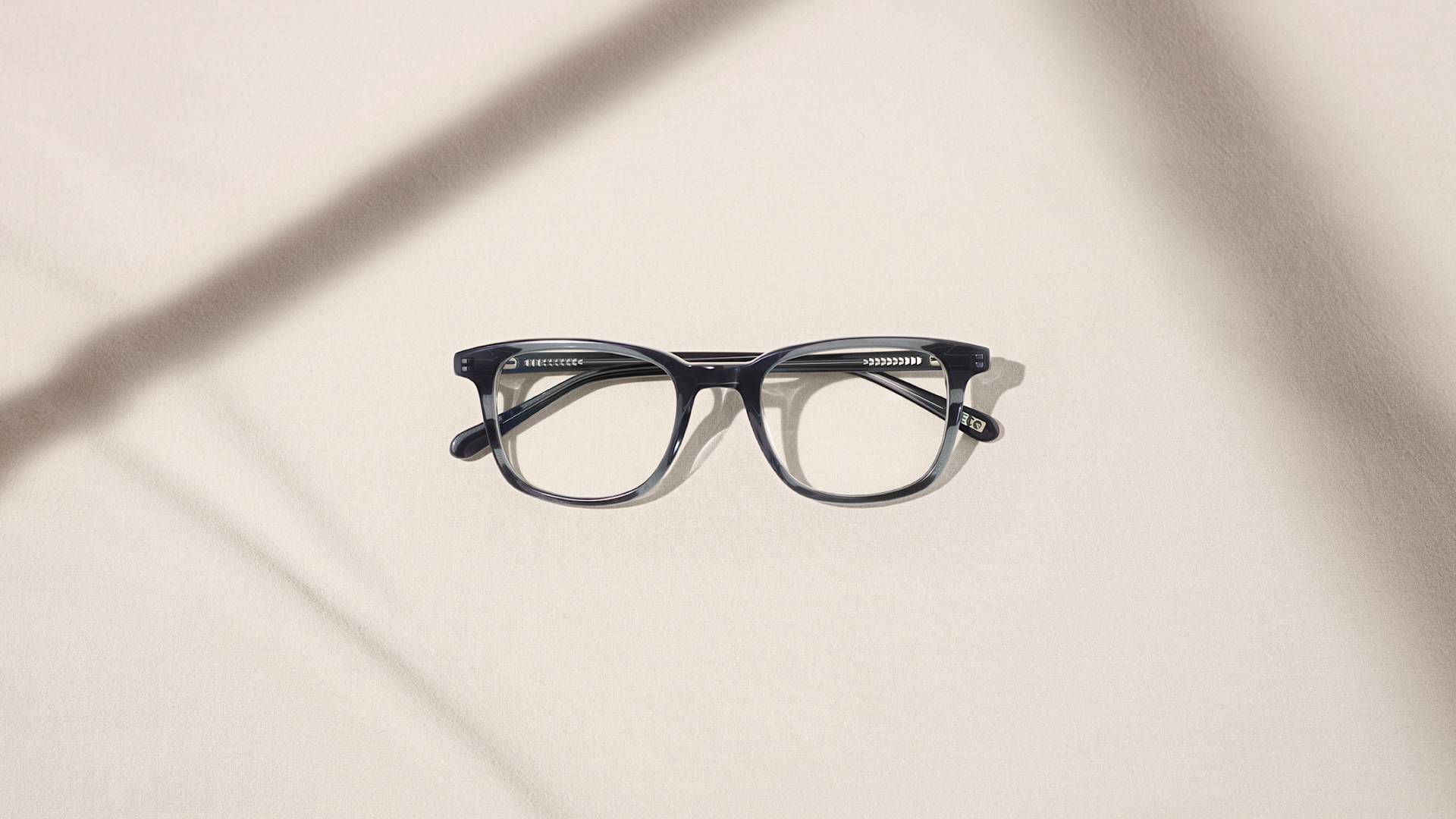 A pair of black-framed eyeglasses lies on a light beige surface, with two soft shadows crossing diagonally across the background.