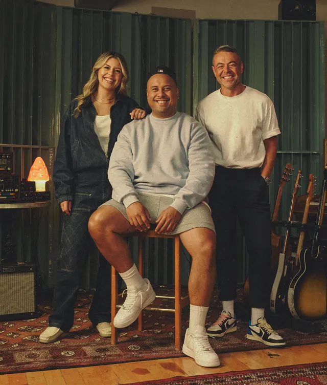 Three people pose and smile in a music studio with guitars, audio equipment, and a Himalayan salt lamp. One person sits on a stool between two standing people. All are dressed casually and appear relaxed and happy.