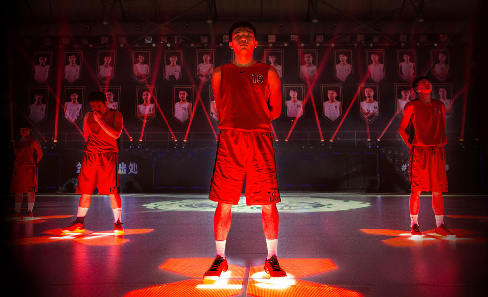 A basketball player in a red uniform stands confidently at center court under dramatic red lighting, with four teammates behind him. Portraits of other players are displayed on the wall in the background.