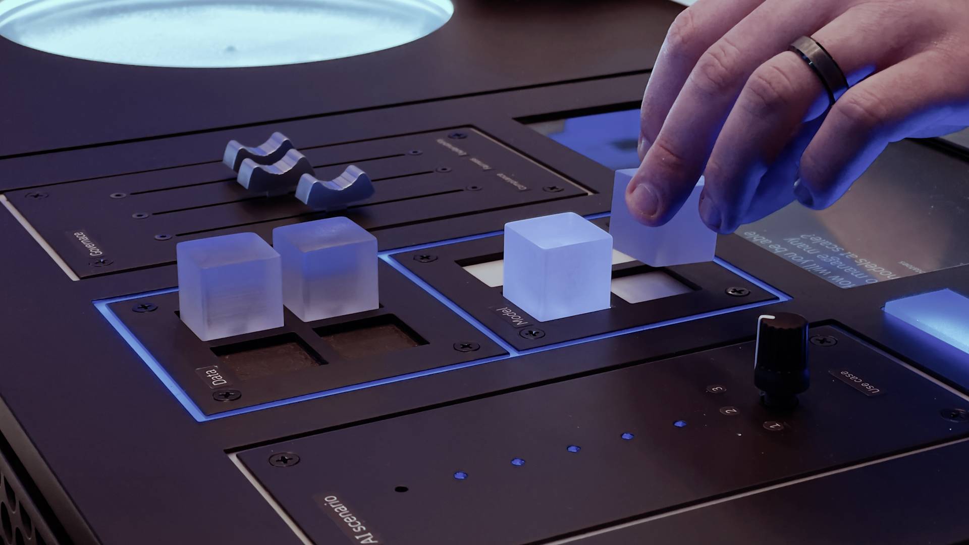 A hand adjusts translucent cubes and sliders on a sleek, modern control panel illuminated with blue light. Other controls, buttons, and a round display are also visible.