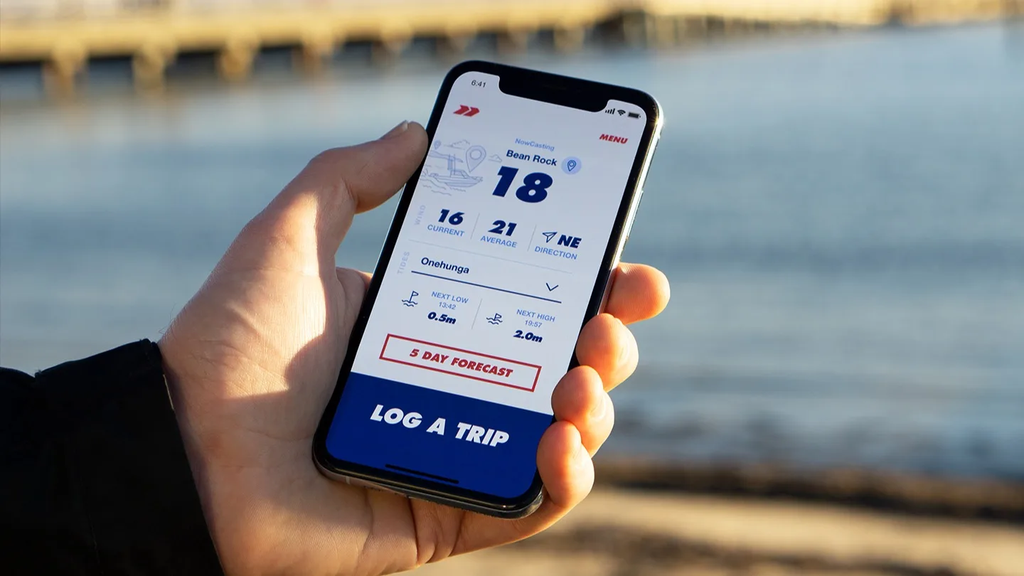 A hand holding a smartphone displaying a boating weather app, showing wind speed, direction, and forecast data, with water and a blurred dock in the background.