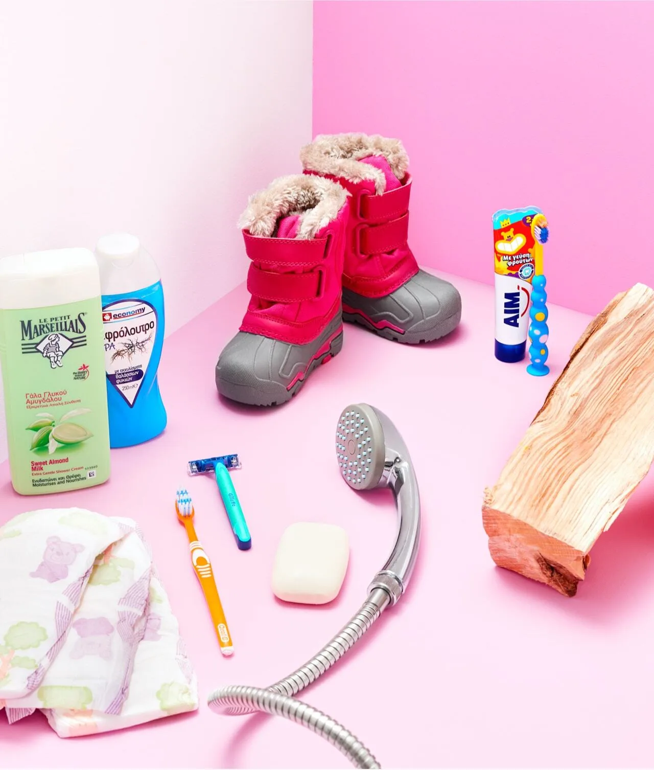 A pair of pink winter boots, soap, shampoo, a toothbrush, toothpaste, baby diaper, a log of wood, and a shower head are arranged on a pink and white background.