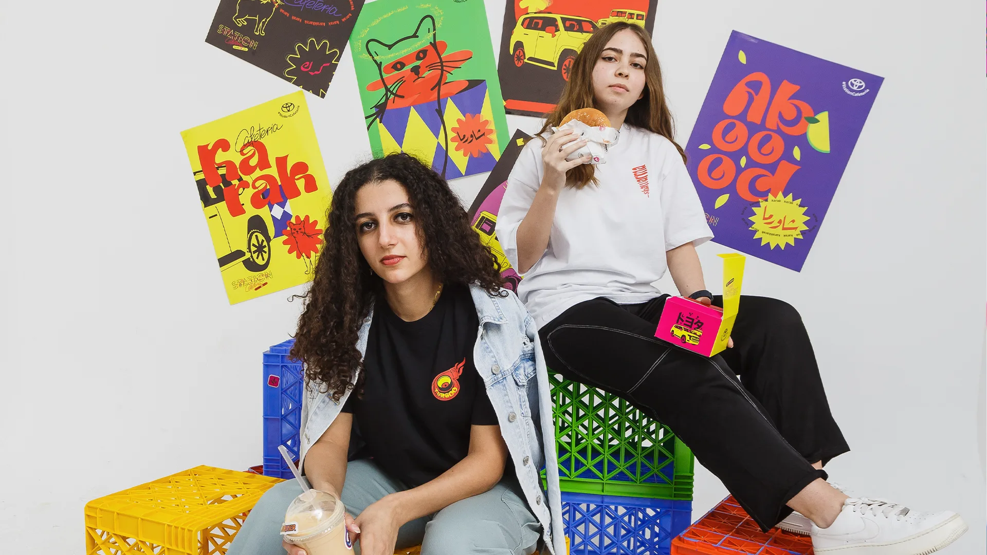 Two young women sit on colorful plastic crates, holding drinks and food. Bright, playful posters featuring bold graphics and text are displayed on the white wall behind them, adding a vibrant and energetic atmosphere.