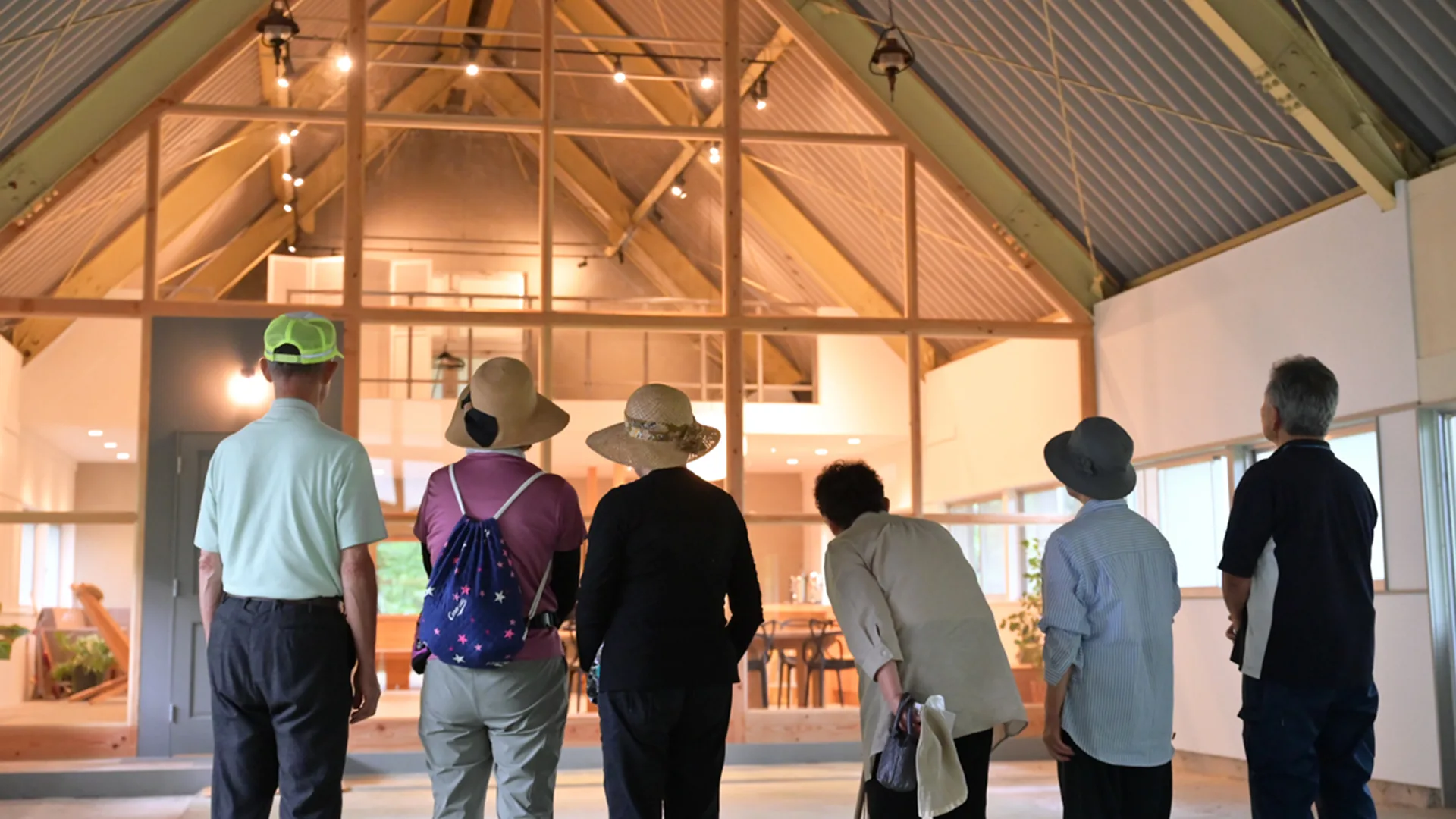 Six older adults, seen from behind, stand inside a large, modern building with high ceilings, looking towards a wooden-framed glass wall and a spacious, warmly lit interior beyond it.