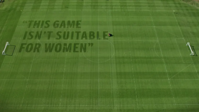 A football field with the text “THIS GAME ISN’T SUITABLE FOR WOMEN” mowed into the grass, viewed from above. A lone person operates a lawnmower at the center of the field.