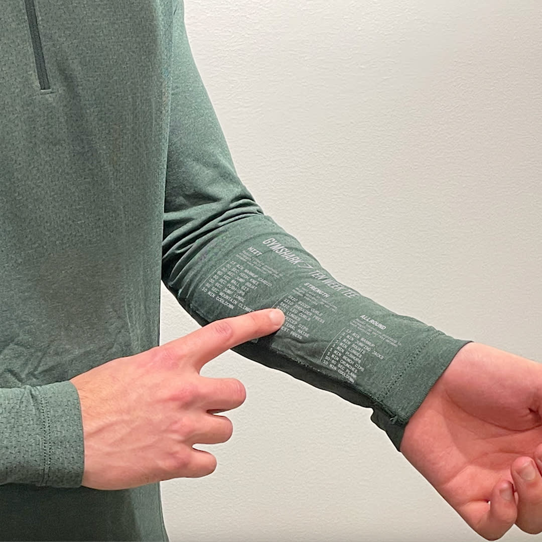 A person wearing a long-sleeve green shirt points to printed text on the inside of their sleeve, with a light-colored wall in the background. Only the persons hands, arm, and torso are visible.