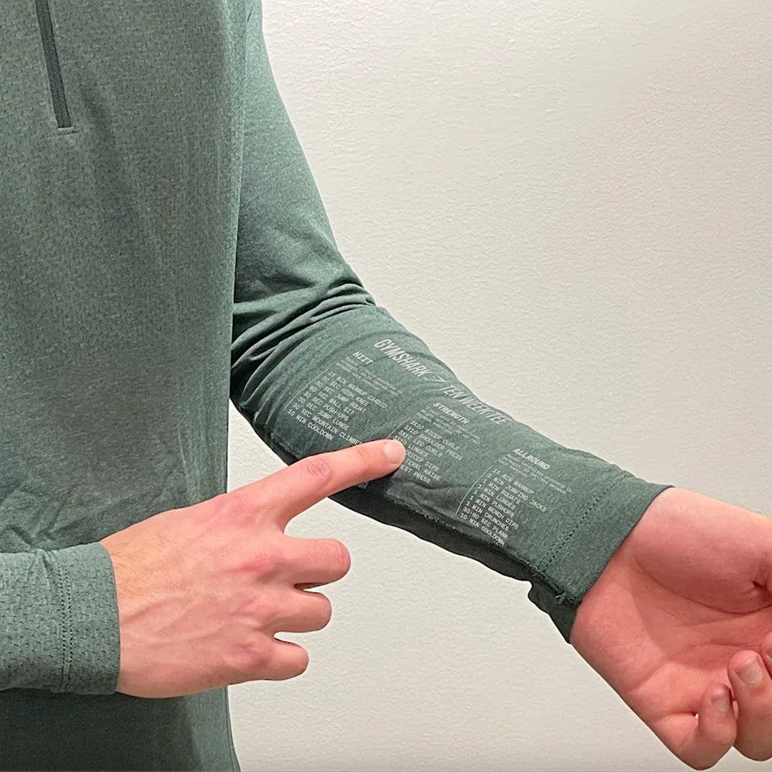 A person wearing a long-sleeve green shirt points to printed text on the inside of their sleeve, with a light-colored wall in the background. Only the persons hands, arm, and torso are visible.