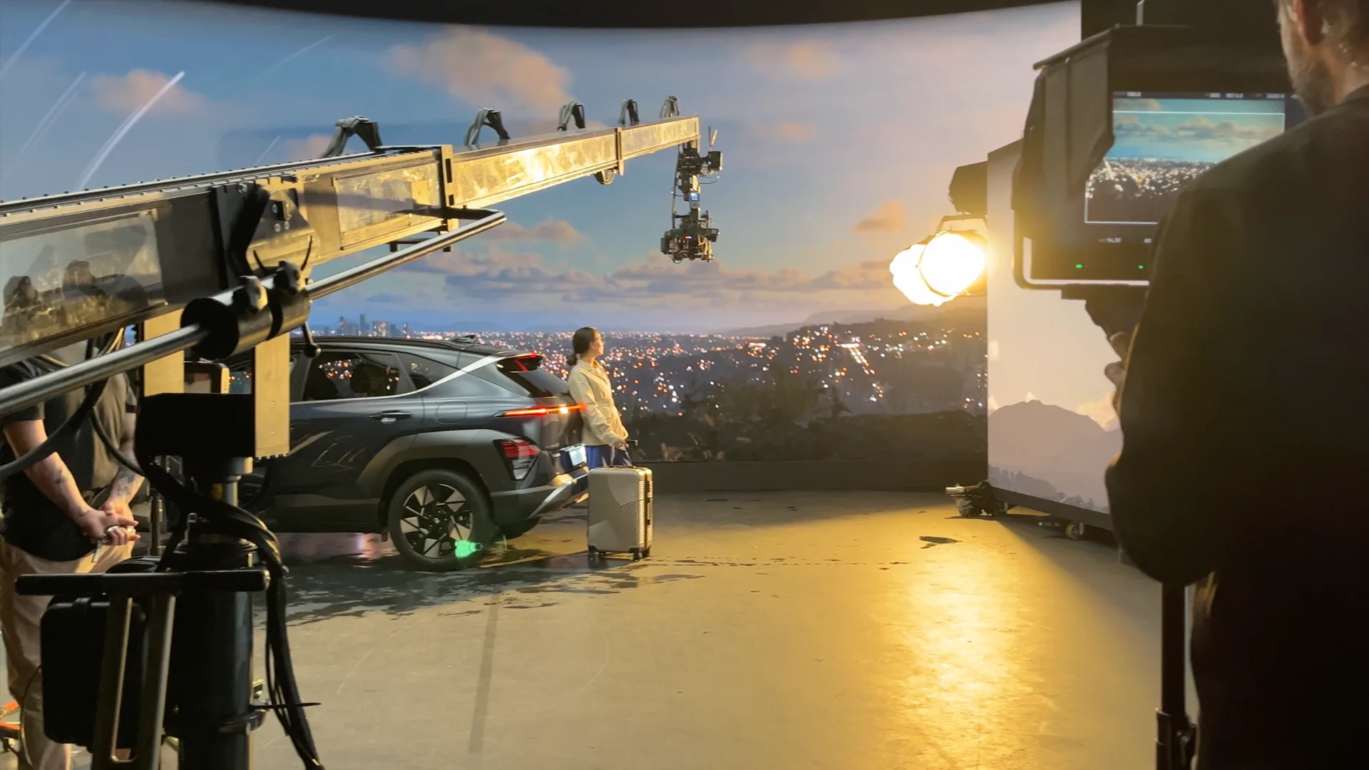 A woman with a suitcase stands next to a car on a film set, surrounded by cameras, lights, and crew. A cityscape is projected in the background, and a monitor displays the scene being filmed.