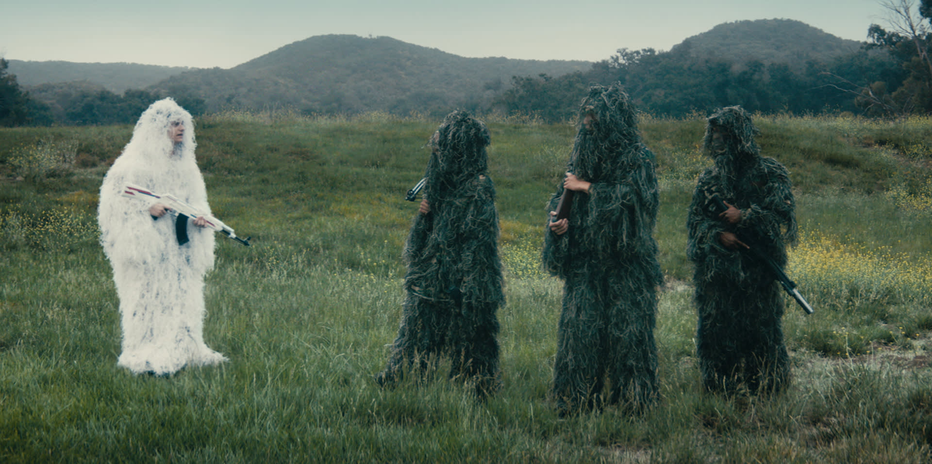 Four people stand in a grassy field; three wear green ghillie suits, while one on the left wears a white ghillie suit and holds a rifle. Hills and trees are in the background under a cloudy sky.