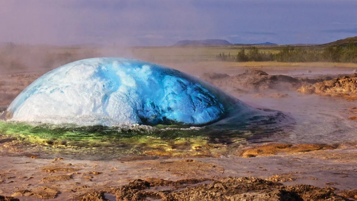 A large, blue, translucent dome of water bulges above a hot spring, surrounded by steam and rocky ground, with green fields and distant hills in the background under a cloudy sky.