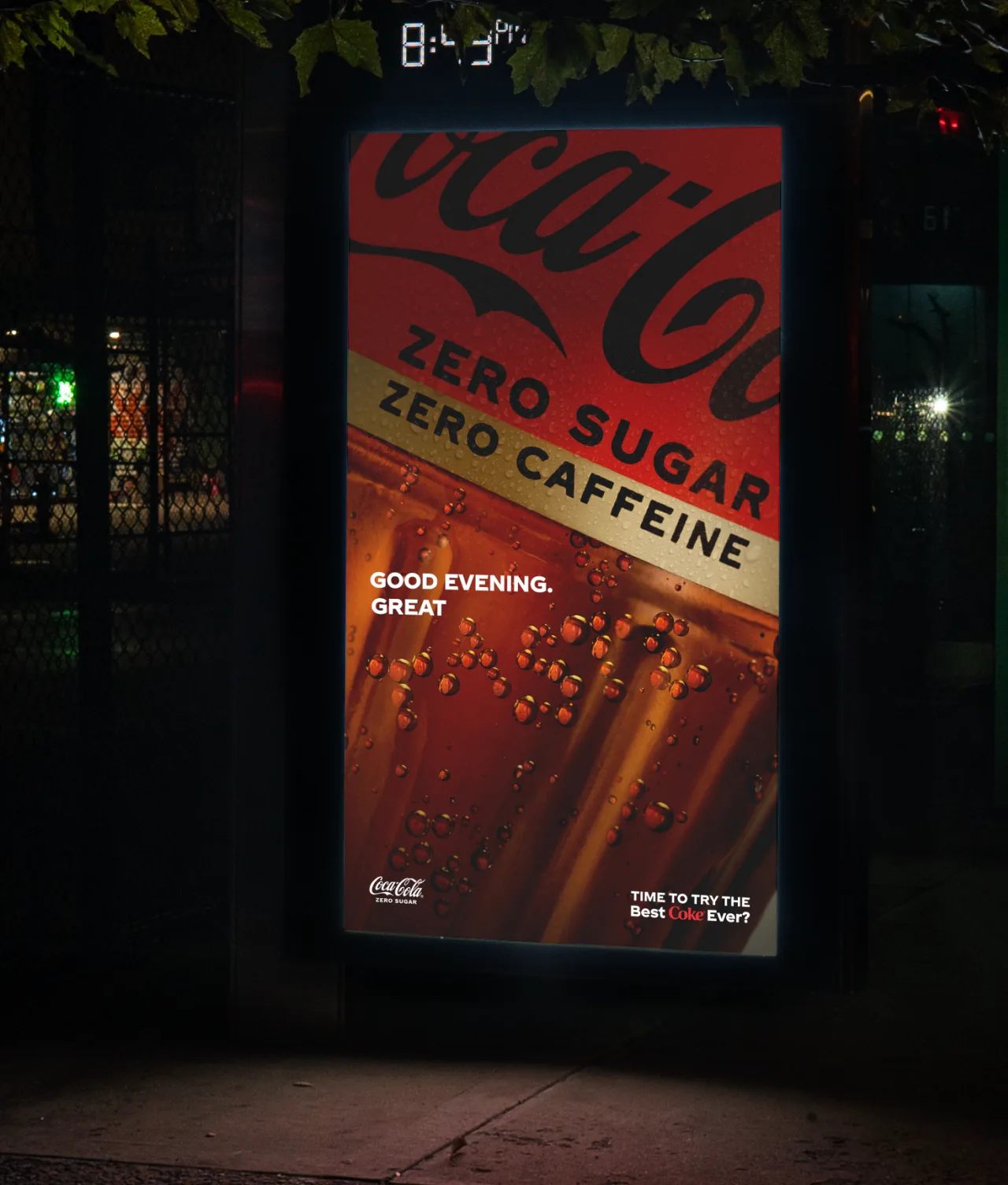 A Coca-Cola Zero Sugar, Zero Caffeine ad displayed at a bus stop at night, featuring a close-up of a fizzy drink, with the text “Good evening. Great” and “Time to try the best Coke ever?”.
