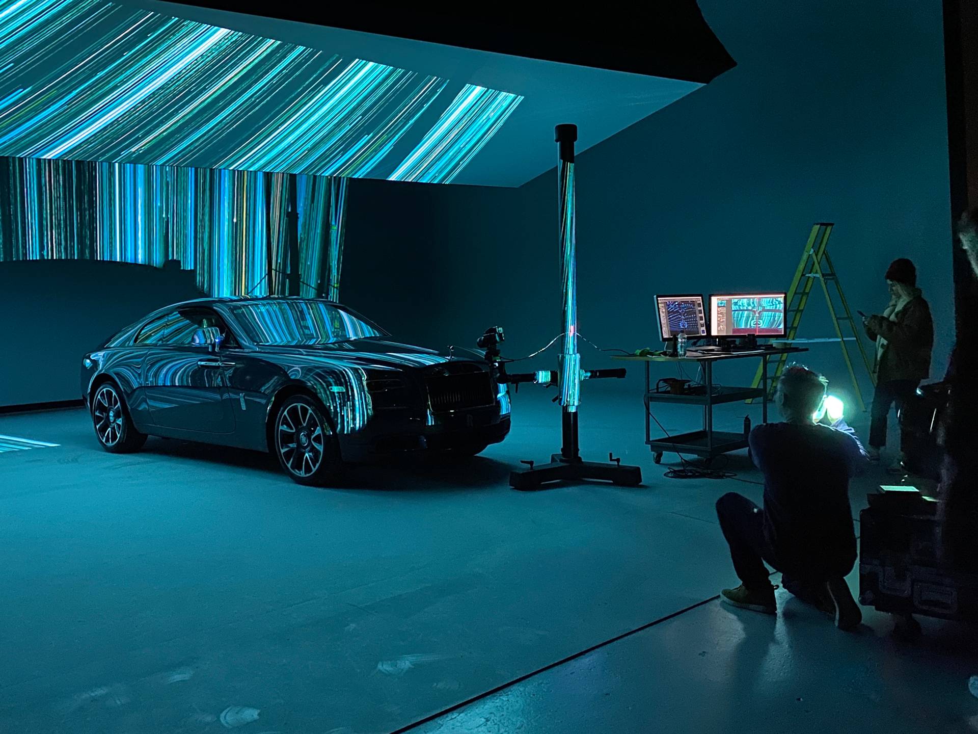 A dark studio with a luxury car under blue lights, projection patterns on the wall and car, a person photographing it, another by computer screens, and a yellow ladder in the background.