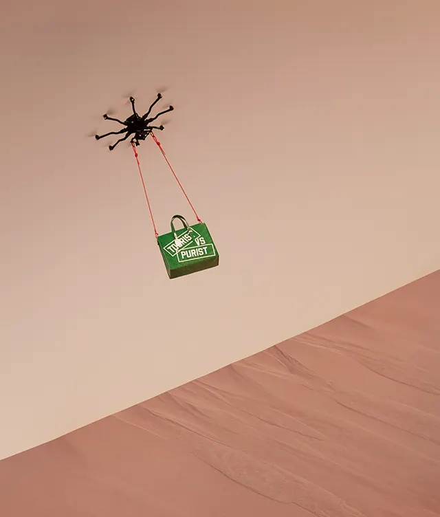 A drone with multiple rotors flies over a sandy desert, carrying a green bag labeled THE FUTURE IS PURIST suspended by two orange straps.