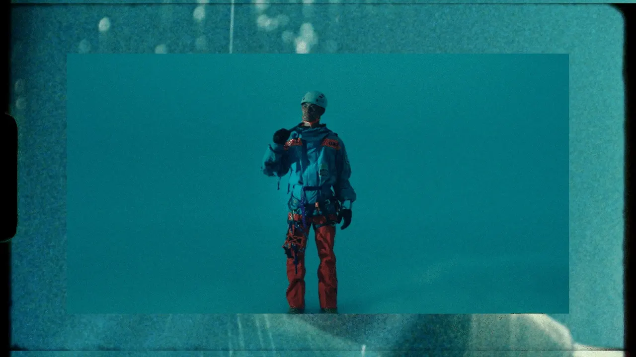 A person wearing a helmet, blue jacket, and red pants stands against a blue background, holding a megaphone. Safety gear and climbing equipment are attached to their belt. The image has a vintage, film-like effect.