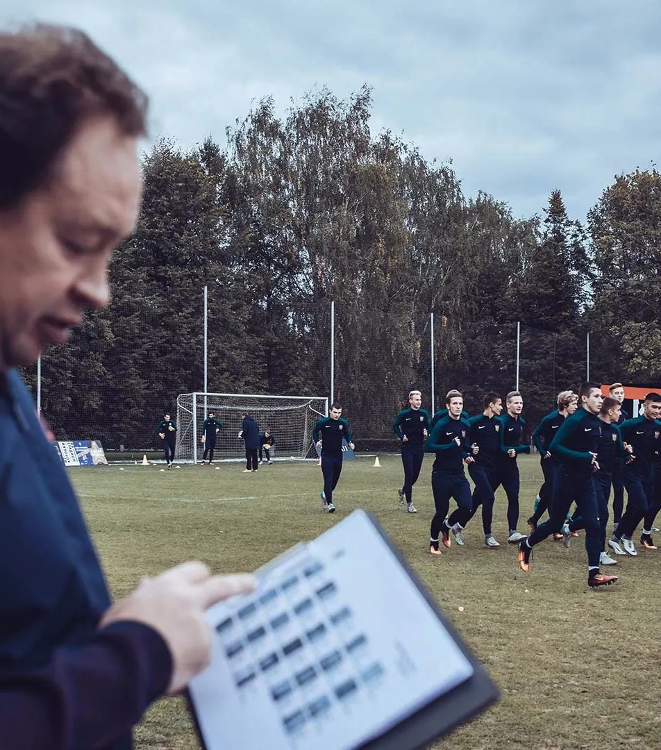 A coach holds a clipboard with notes in the foreground while a group of soccer players in dark athletic gear jog together on a grassy field near a goalpost, surrounded by trees under a cloudy sky.