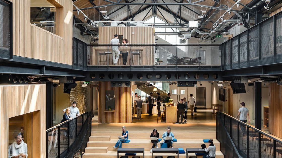 Modern open-plan office space with high ceilings, wooden accents, stairs, seating areas, and people working and socializing on both levels under a glass roof.