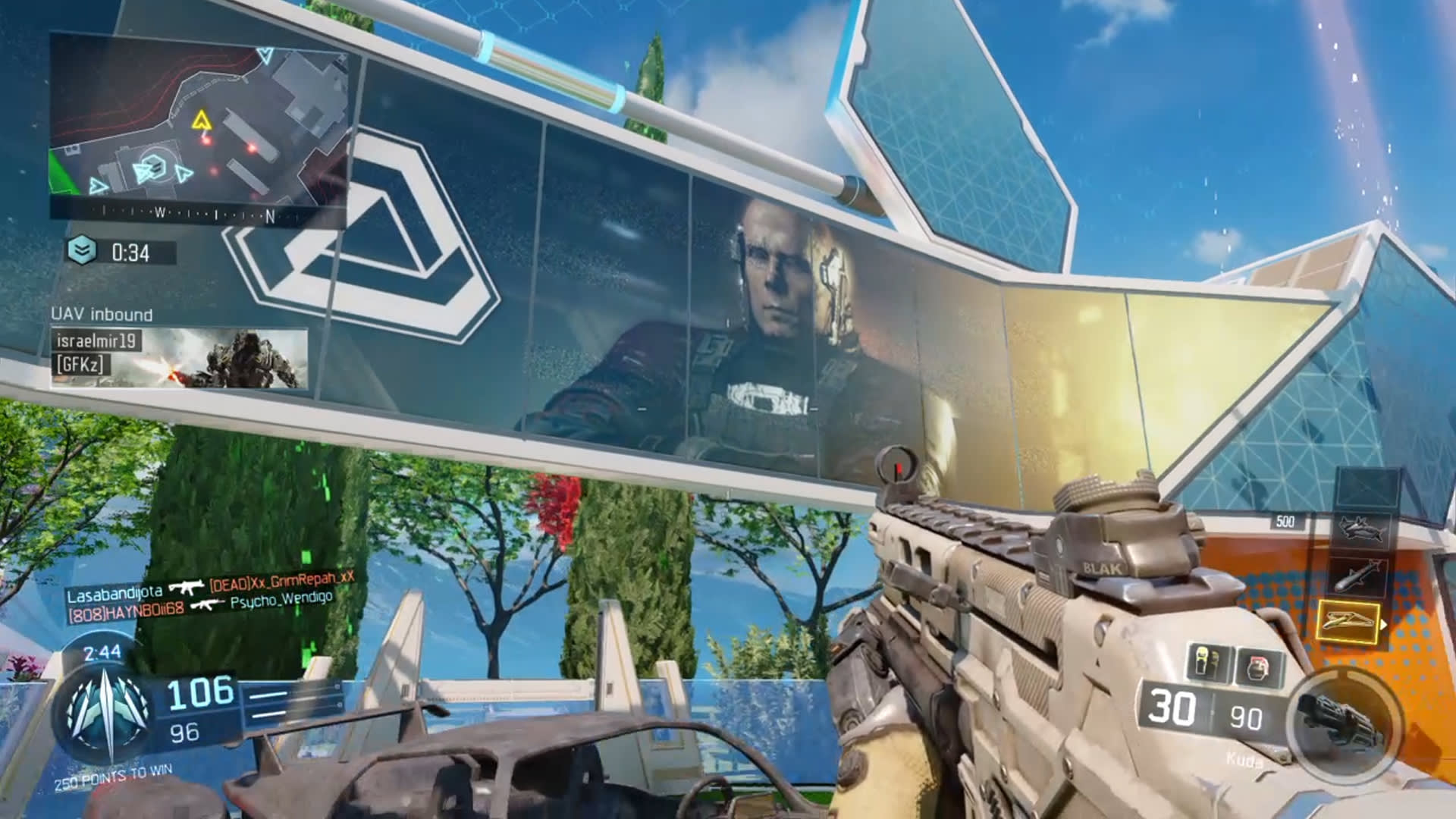A first-person shooter video game scene shows a player holding a futuristic rifle, aiming at a large outdoor digital billboard displaying a soldiers face. The setting is bright with blue skies, trees, and modern structures.