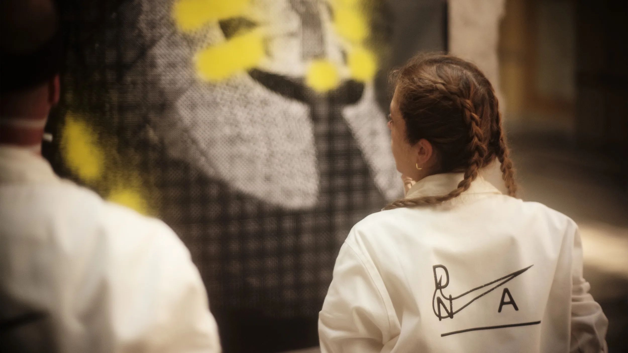 A person with braided hair wearing a white jacket with a unique logo stands indoors, facing a large canvas with abstract black, white, and yellow patterns. Another person in a similar jacket is partially visible.