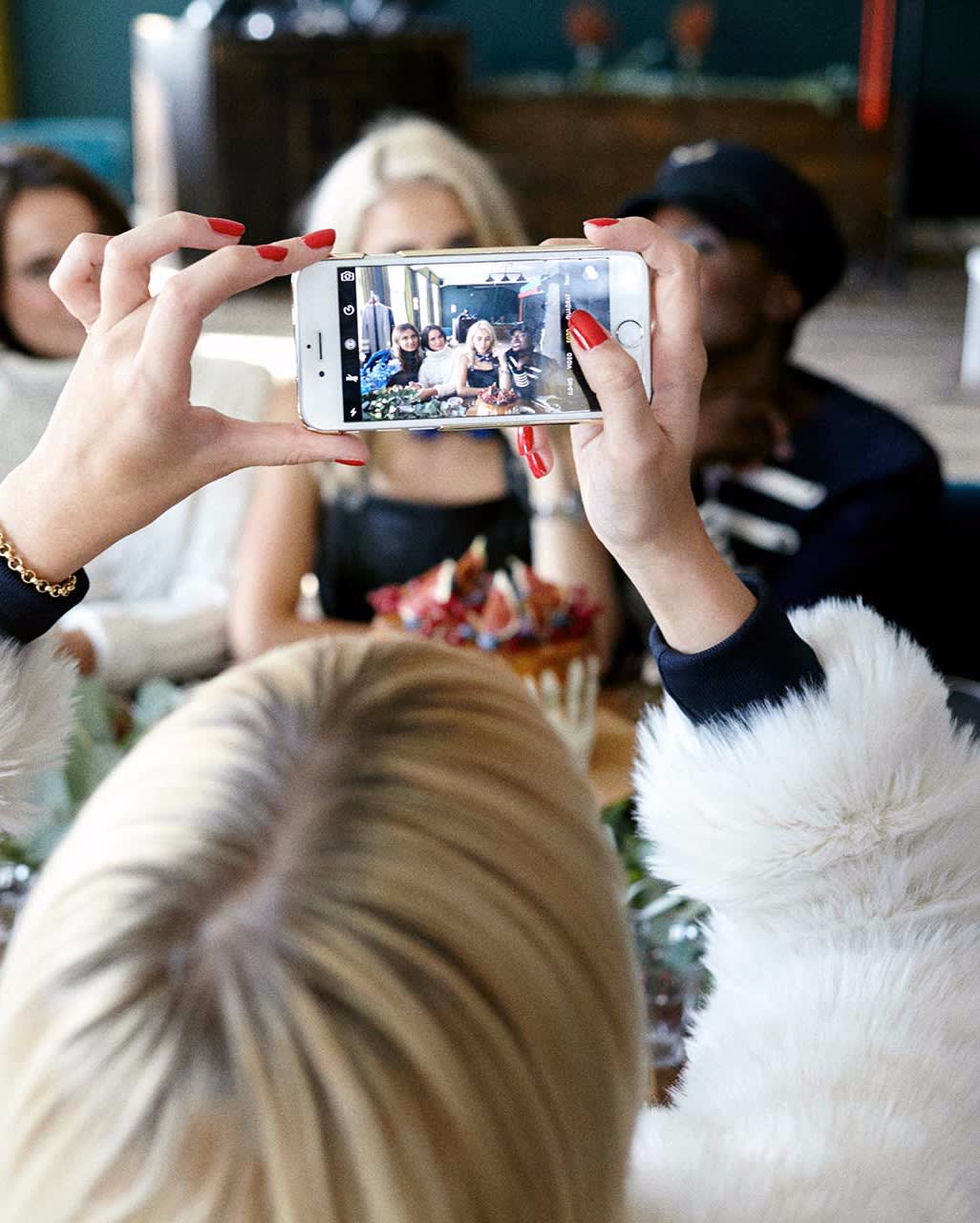 A person with blonde hair and red nails takes a photo with a smartphone of a group of friends sitting together at a table, all smiling and posing for the picture.