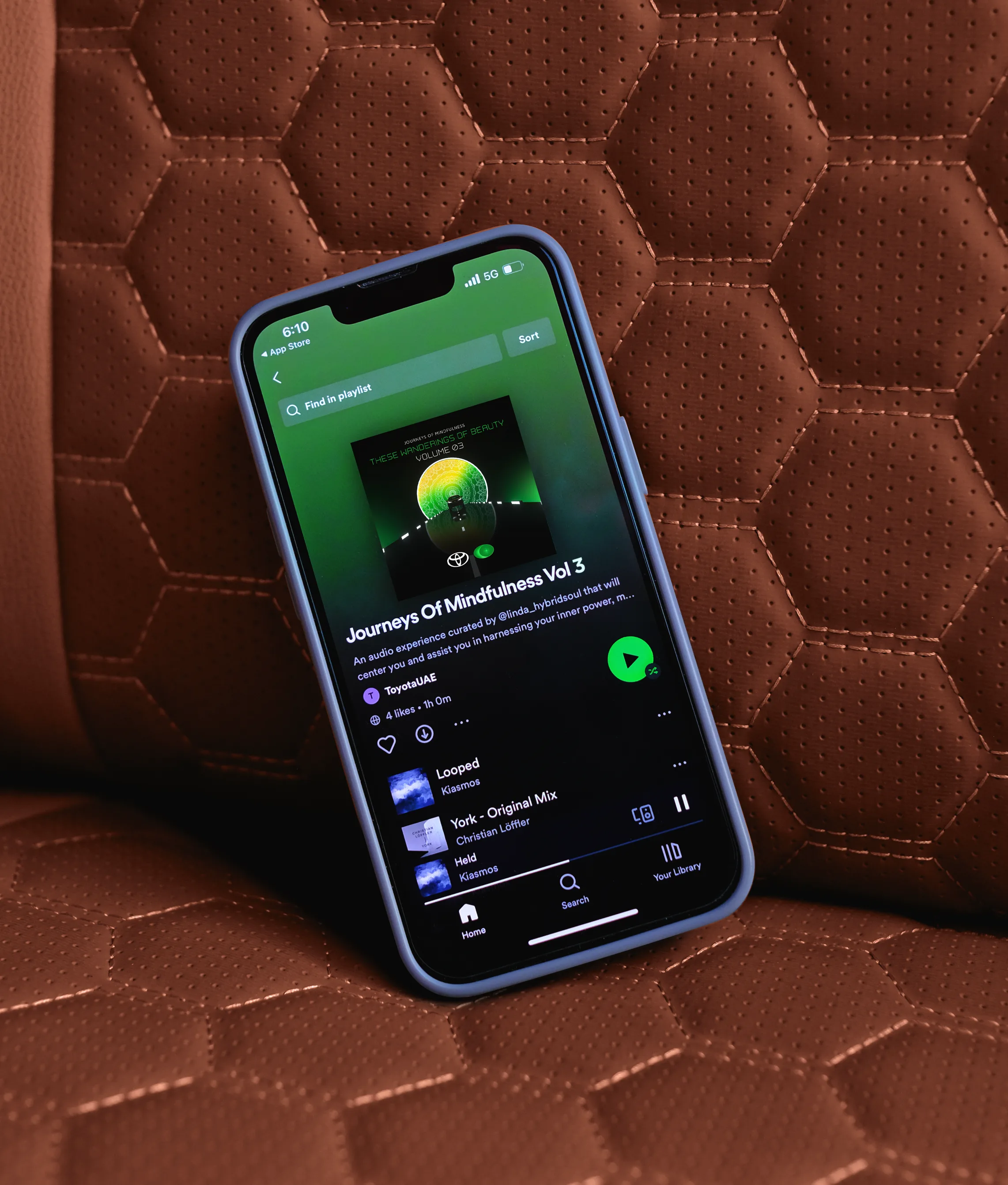 A smartphone displaying a Spotify playlist called Journeys Of Mindfulness Vol 3 rests on a brown, textured car seat with a hexagonal pattern.