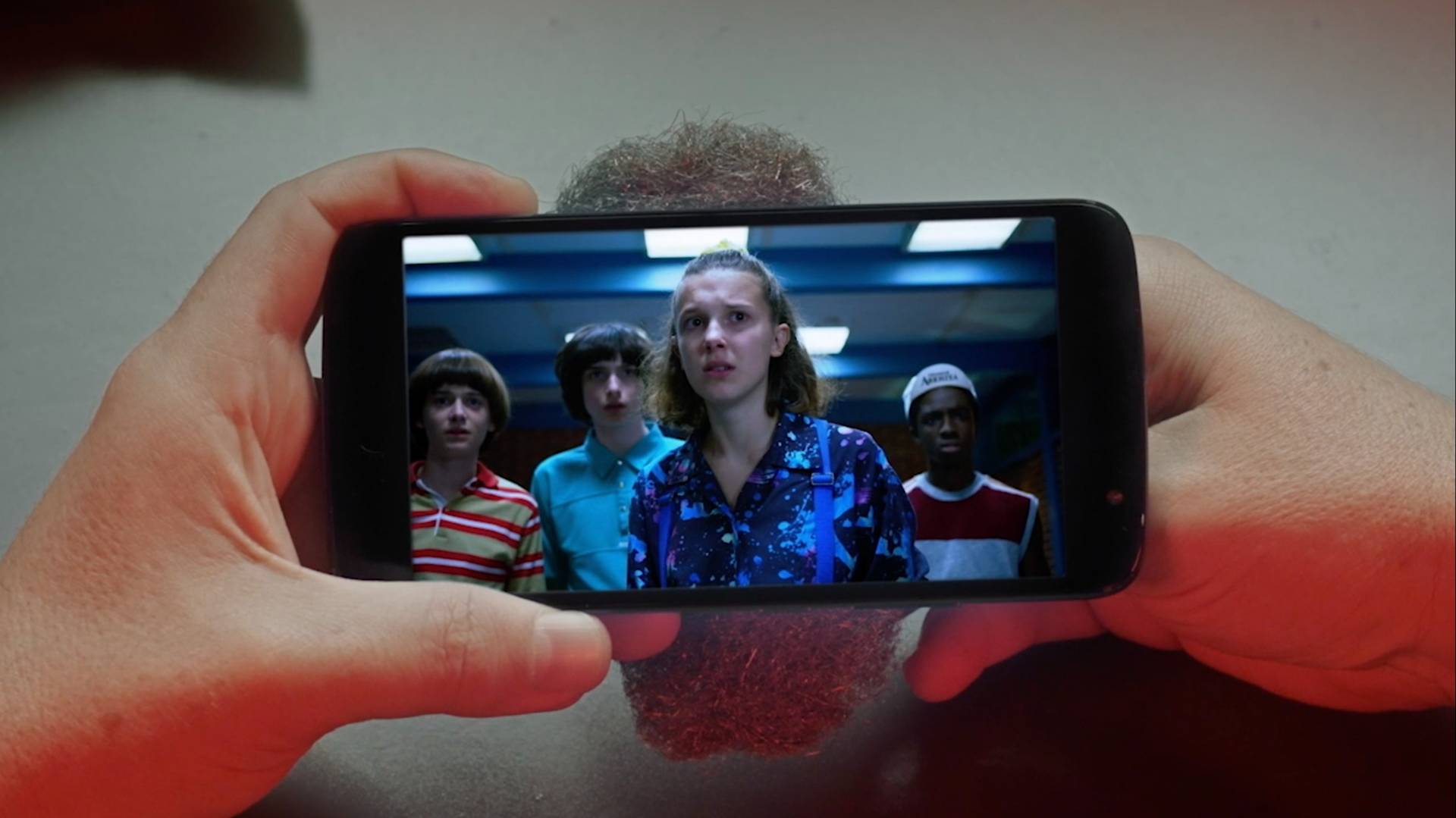 A person holds a smartphone displaying a scene from a TV show with four teenagers looking ahead with serious expressions. The phone is held with both hands over a table.