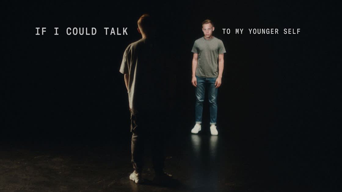 A man stands facing his younger self in a dimly lit space, with the text IF I COULD TALK TO MY YOUNGER SELF above them. Both figures appear serious, the younger self illuminated more brightly.