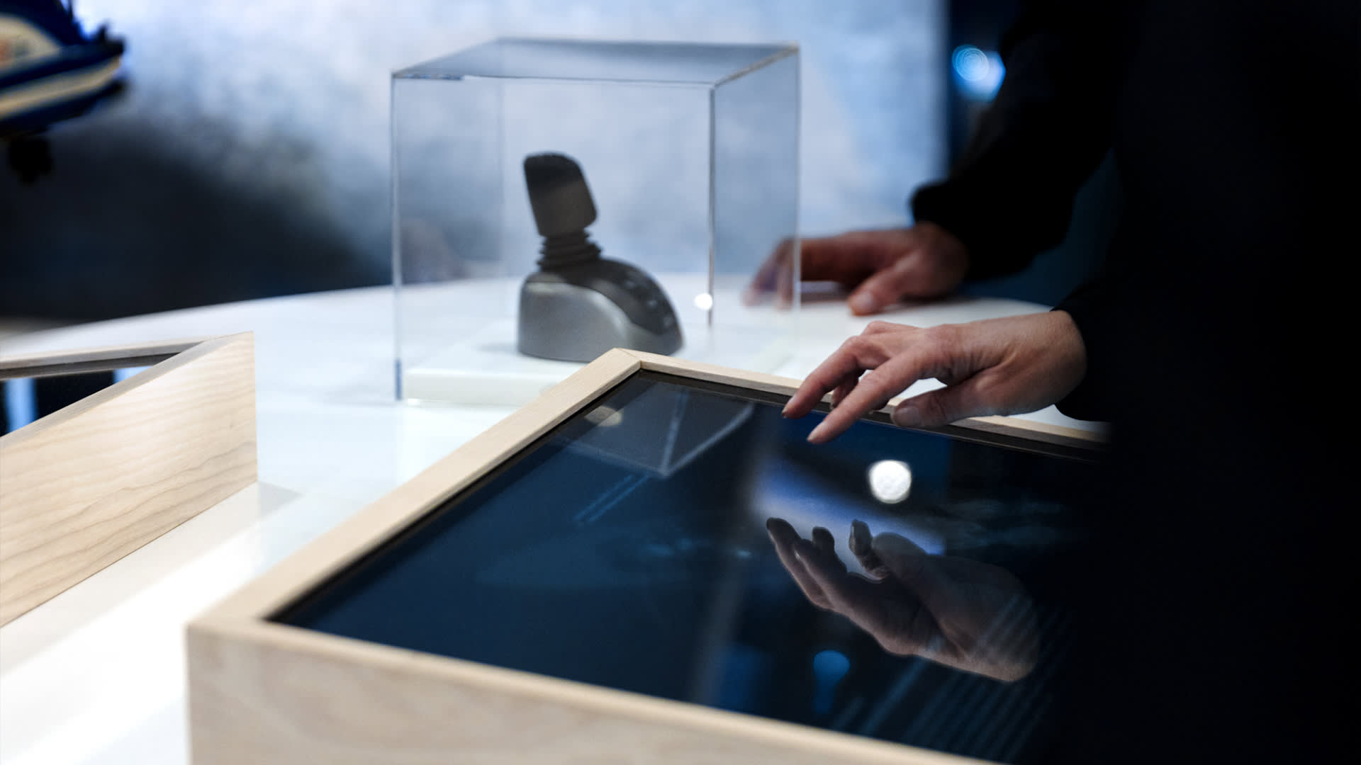 A person interacts with a touchscreen display in a wooden frame, while another hand rests nearby. In the background, a small object is displayed inside a transparent case.