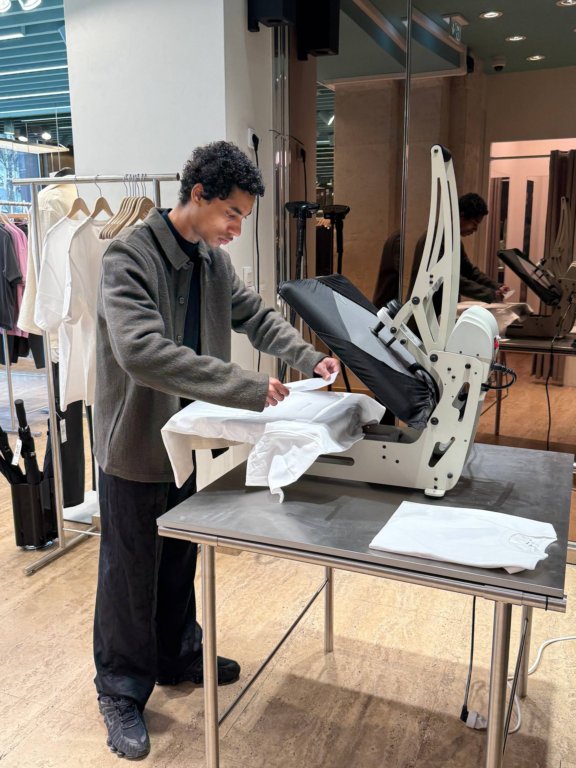 A person stands at a table using a heat press machine to print a design onto a white shirt in a modern clothing store. Shirts on hangers and large windows are visible in the background.