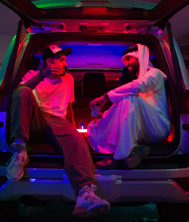 Two men sit in the back of an SUV lit with colorful neon lights. One wears casual clothes and a cap, while the other wears traditional Middle Eastern attire. They are smiling and engaged in conversation.