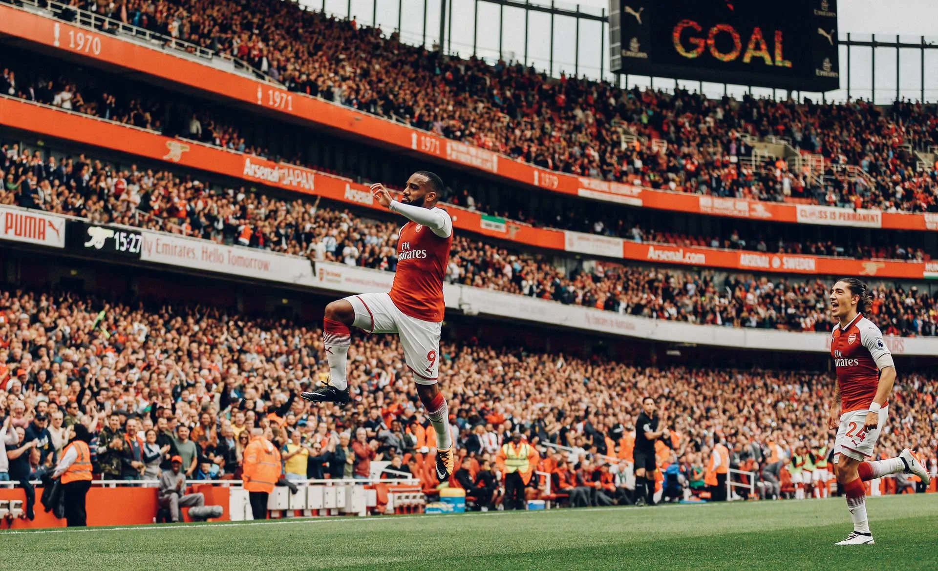 A soccer player in an Arsenal jersey jumps in celebration on the field, while another player approaches. A large crowd of cheering fans fills the stadium, and a “GOAL” sign is visible in the background.