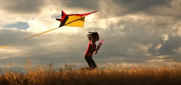 A child runs through a grassy field at sunset, holding a string and flying a yellow and red kite against a dramatic, cloudy sky.