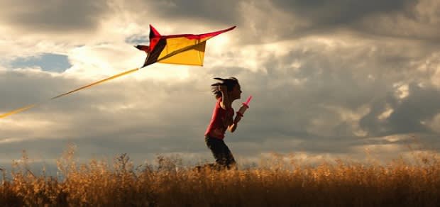 A child runs through a grassy field at sunset, holding a string and flying a yellow and red kite against a dramatic, cloudy sky.