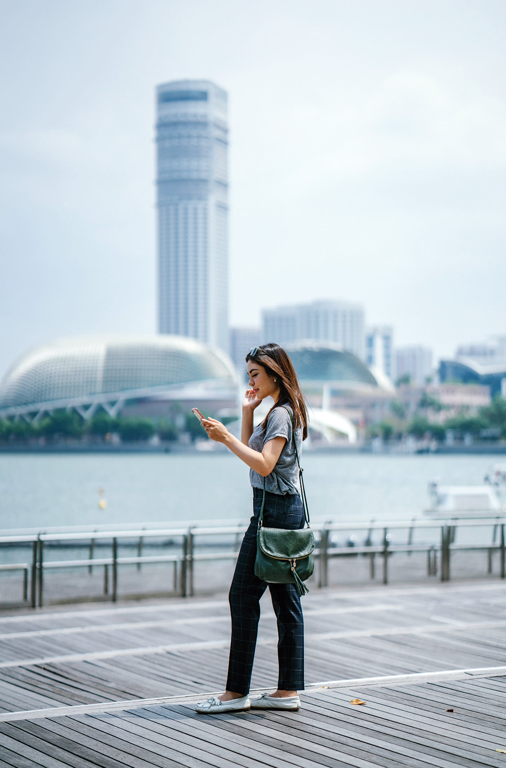 A woman walks along a wooden boardwalk near water, looking at her phone. Modern buildings and a dome-shaped structure are visible in the background under a cloudy sky.