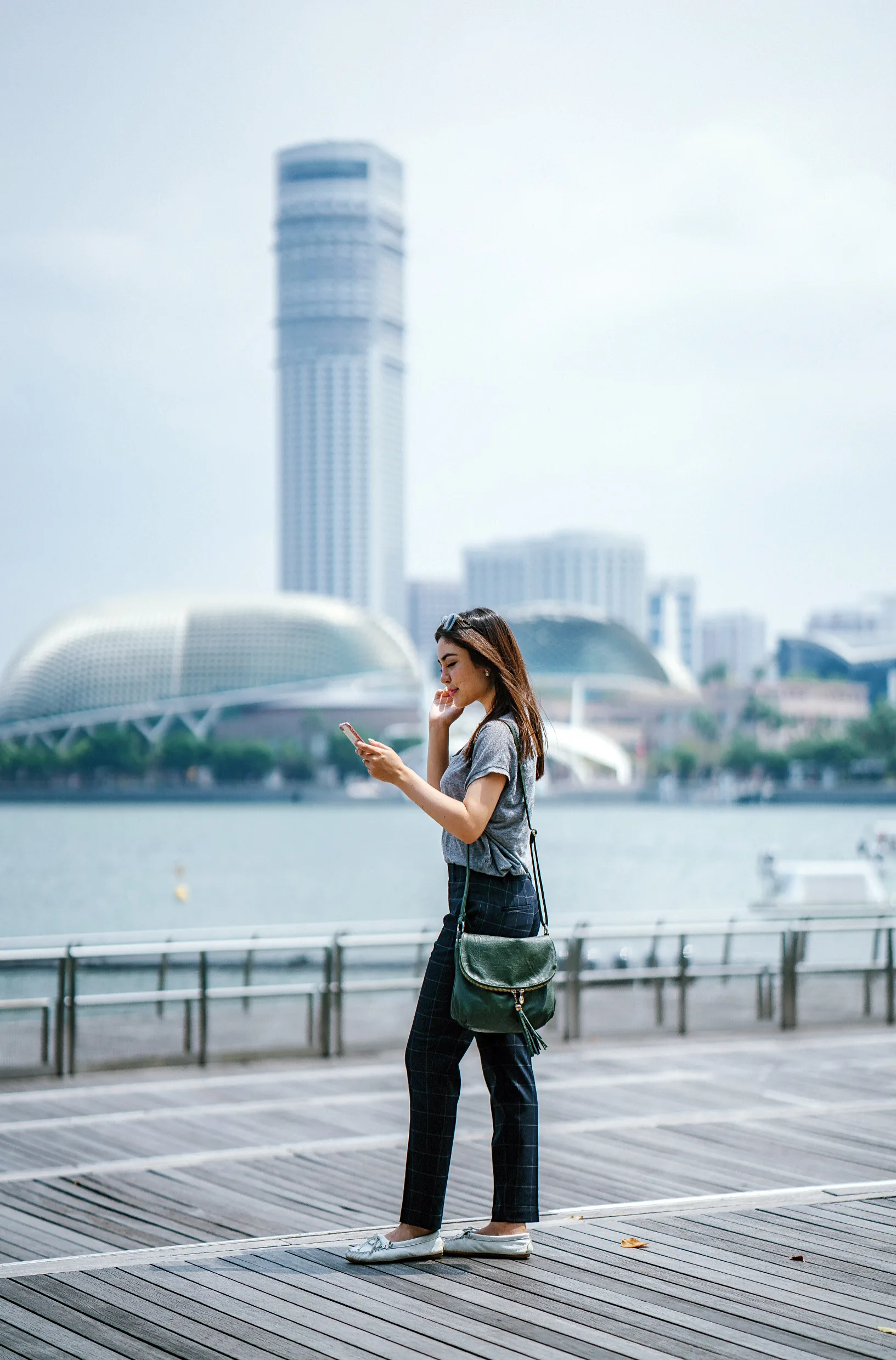 A woman walks along a wooden boardwalk near water, looking at her phone. Modern buildings and a dome-shaped structure are visible in the background under a cloudy sky.