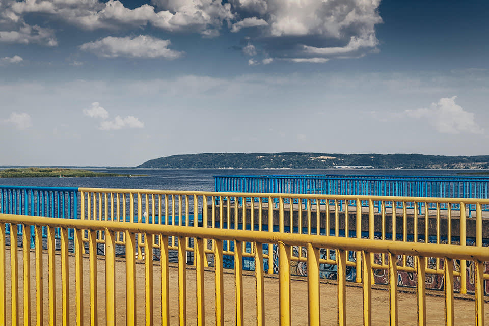 Yellow and blue metal railings line a walkway near a body of water, with distant hills and a partly cloudy sky in the background. The colors create a striking, layered visual effect.