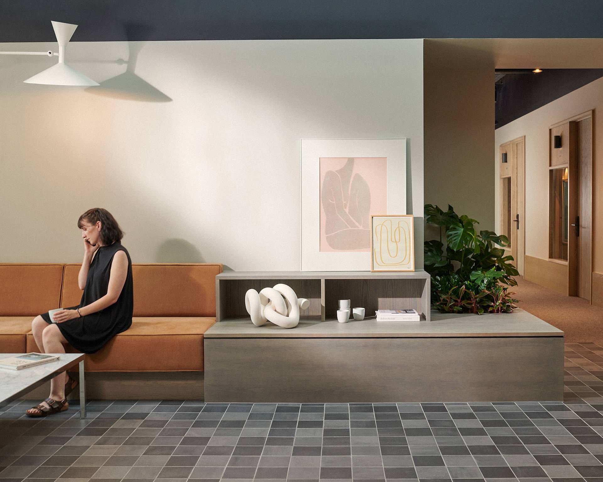 A woman in a black dress sits on an orange bench in a modern, minimalist lobby with abstract art, plants, decorative objects, and a tiled floor. She appears to be talking on the phone.