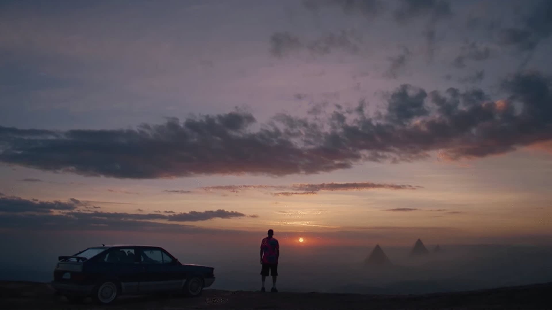 A person stands next to a parked car, gazing at a colorful sunset with the silhouettes of pyramids visible in the distance under a dramatic, cloud-filled sky.
