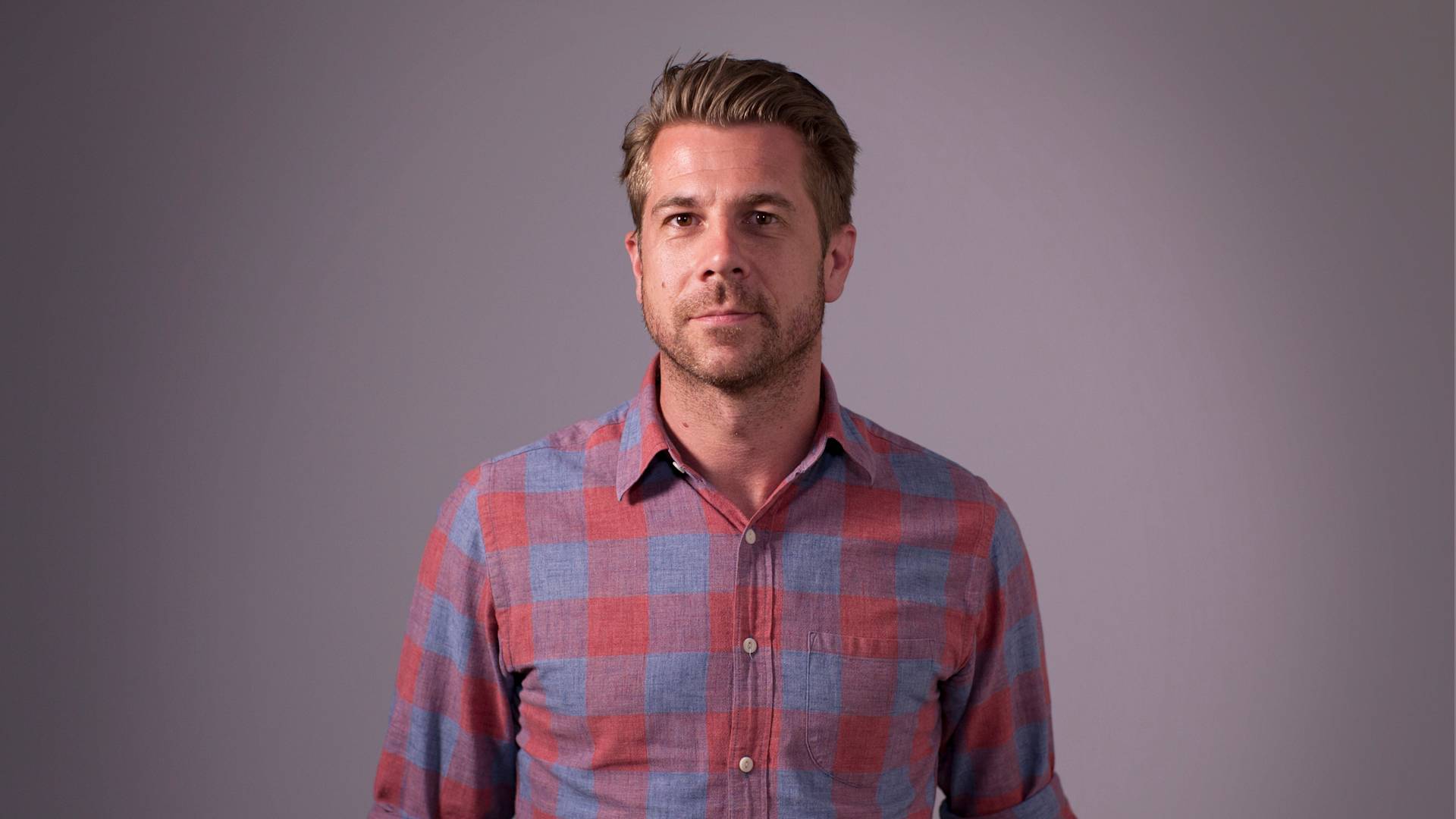 A man with short, light brown hair and stubble is wearing a red and blue plaid button-up shirt. He stands against a plain, light gray background and looks directly at the camera with a neutral expression.